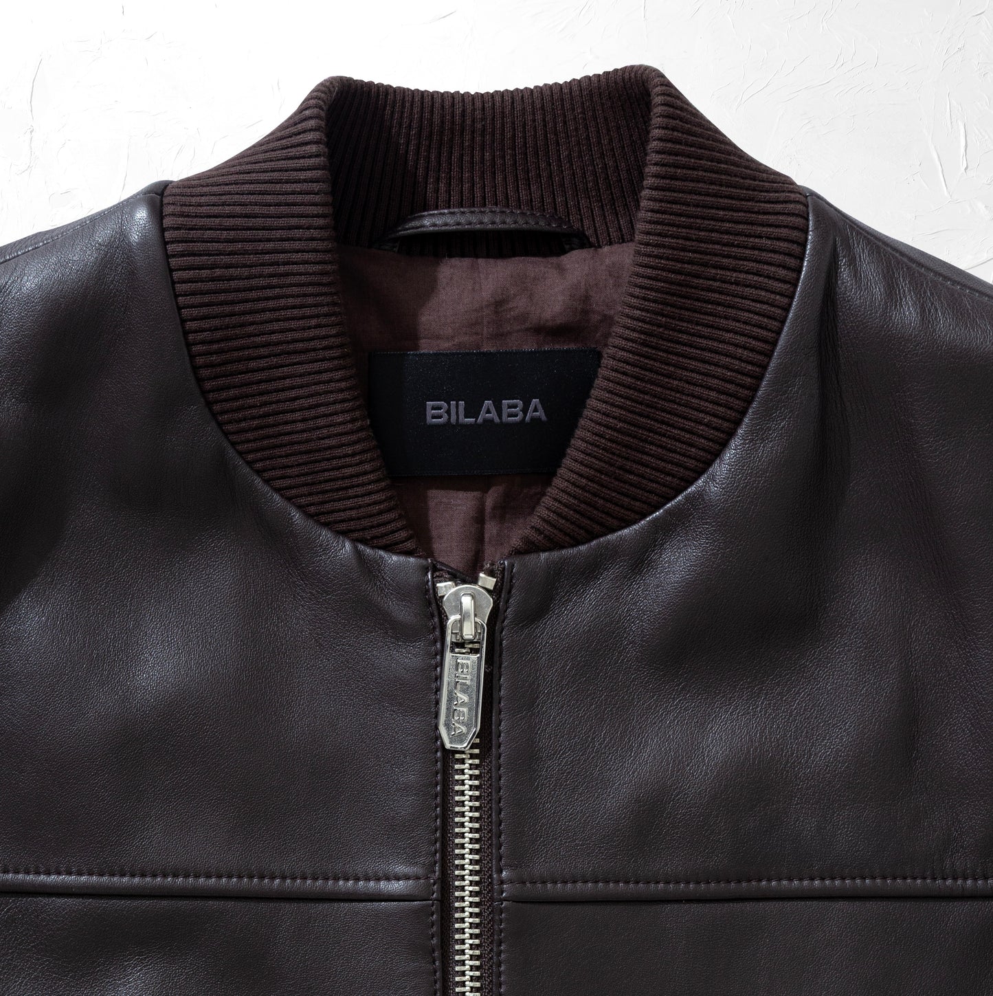 Close-up of a brown bomber leather jacket with a visible brand label 'BILABA' on a white background