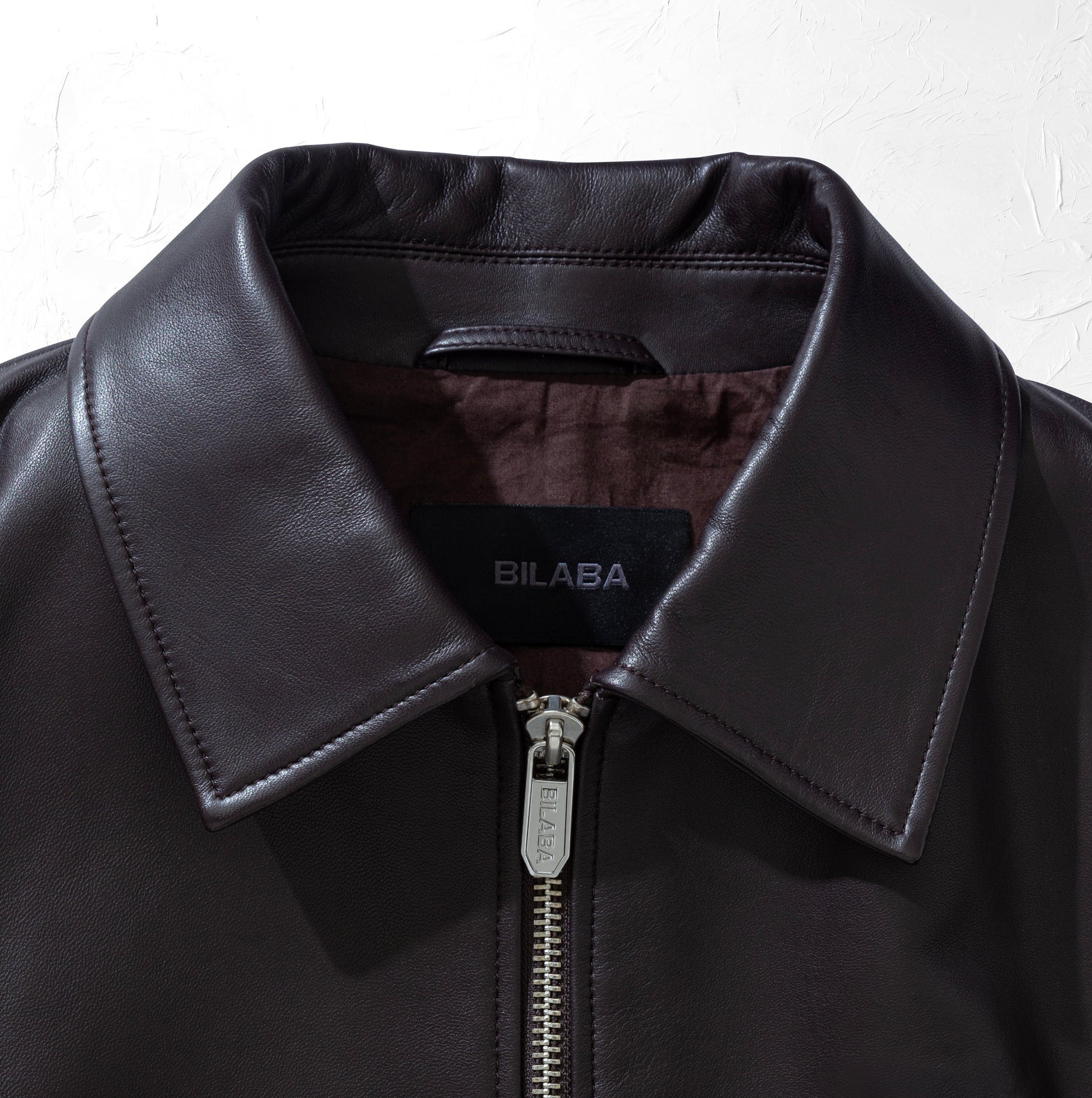 Close-up of a brown leather bomber jacket with a visible brand logo on a white background
