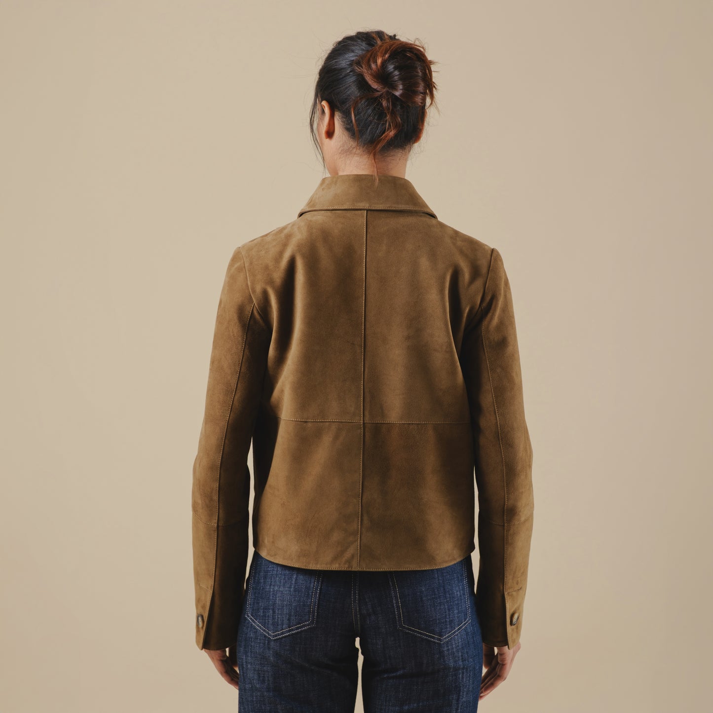 Person wearing a brown suede jacket and blue jeans against a beige background