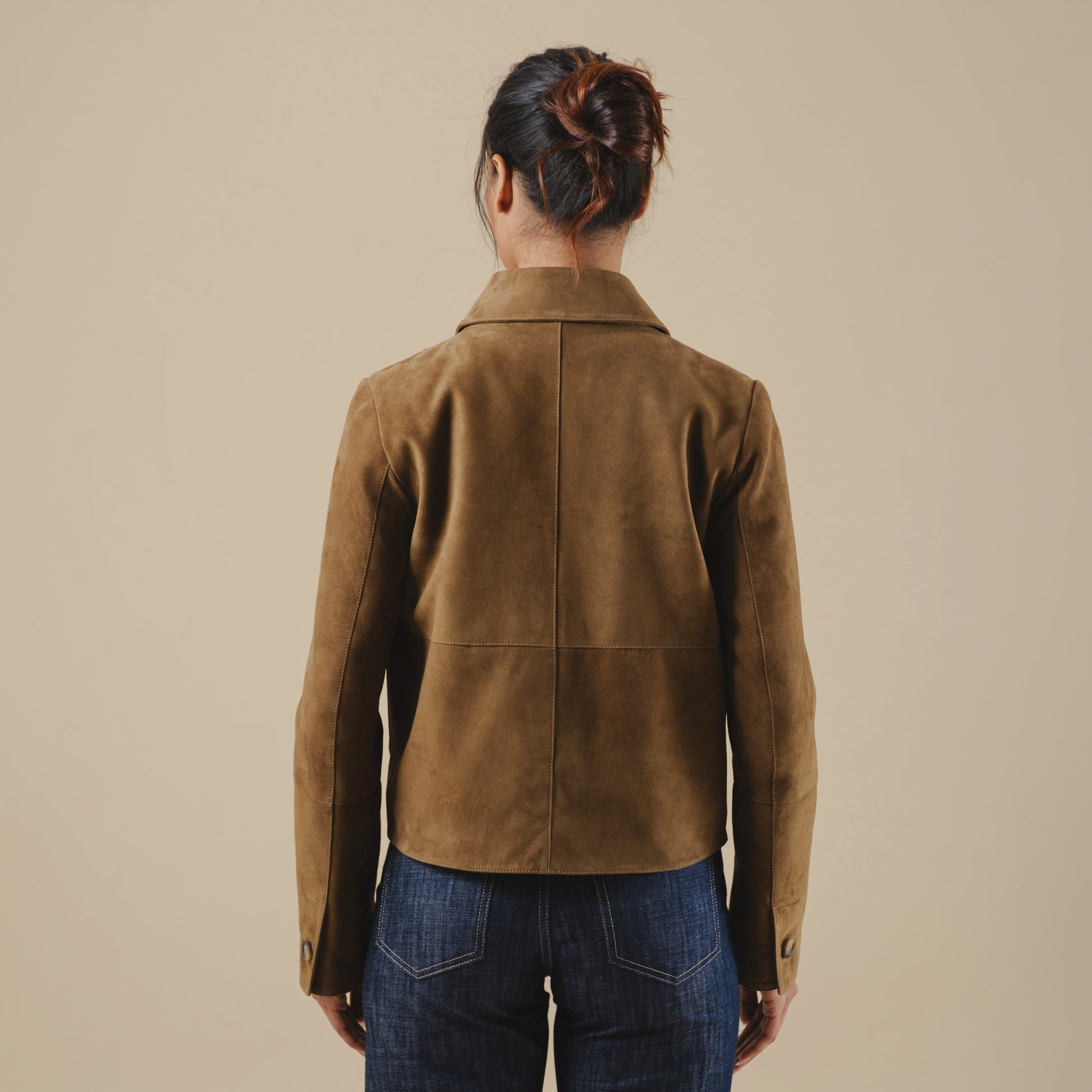 Person wearing a brown suede jacket and blue jeans against a beige background