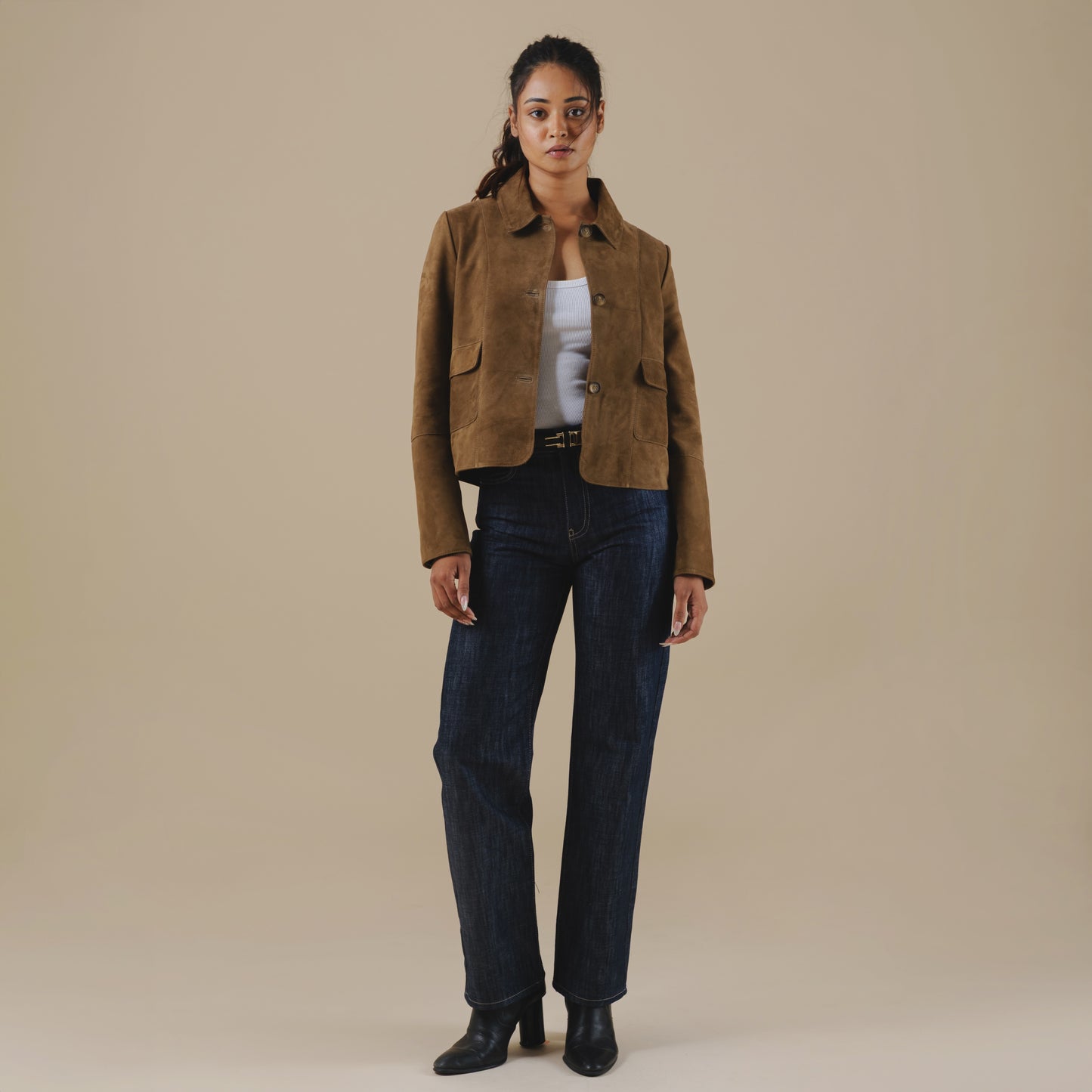 Woman wearing a brown suede jacket and dark jeans against a beige background