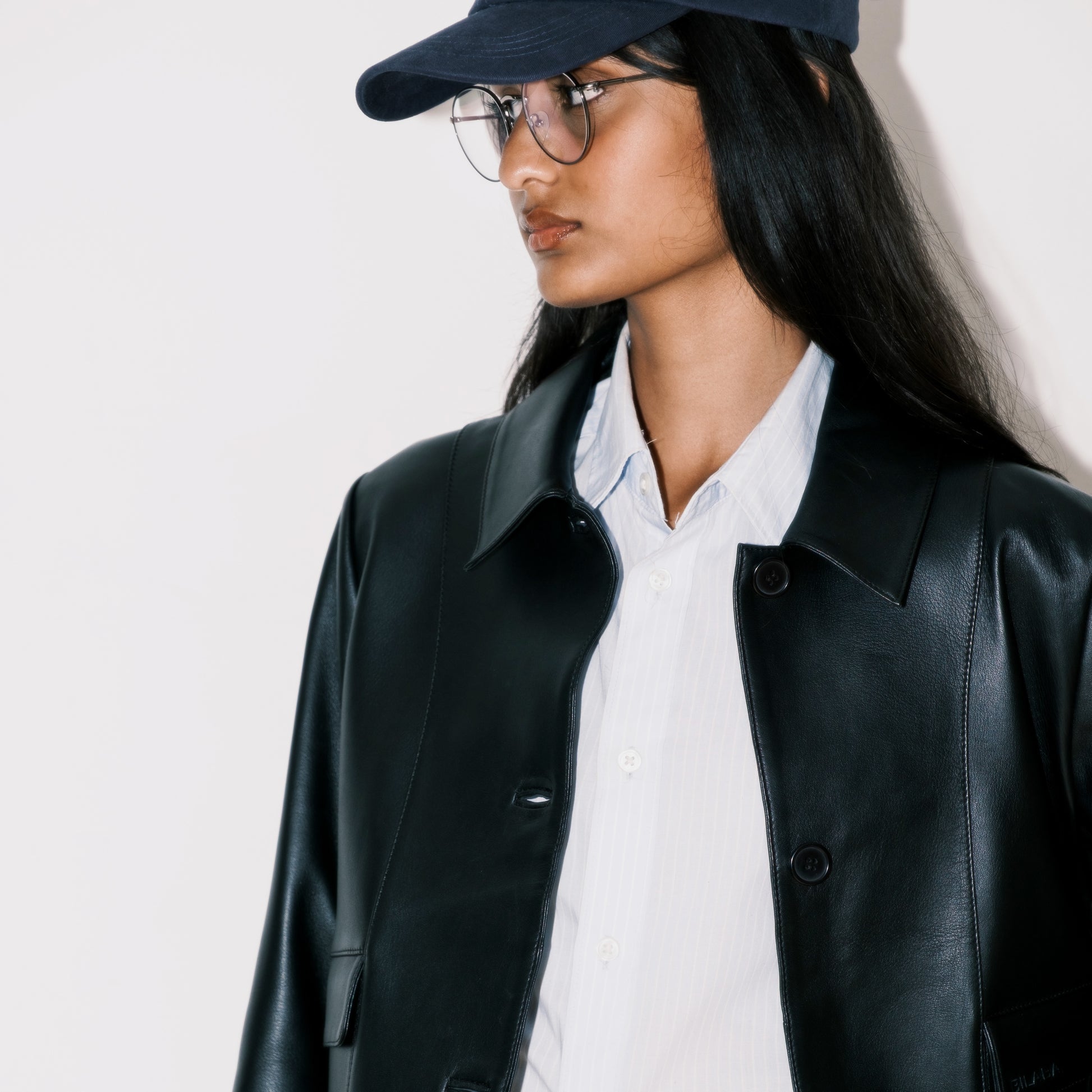Woman wearing a black collared button leather jacket, white shirt, and navy cap on a light background