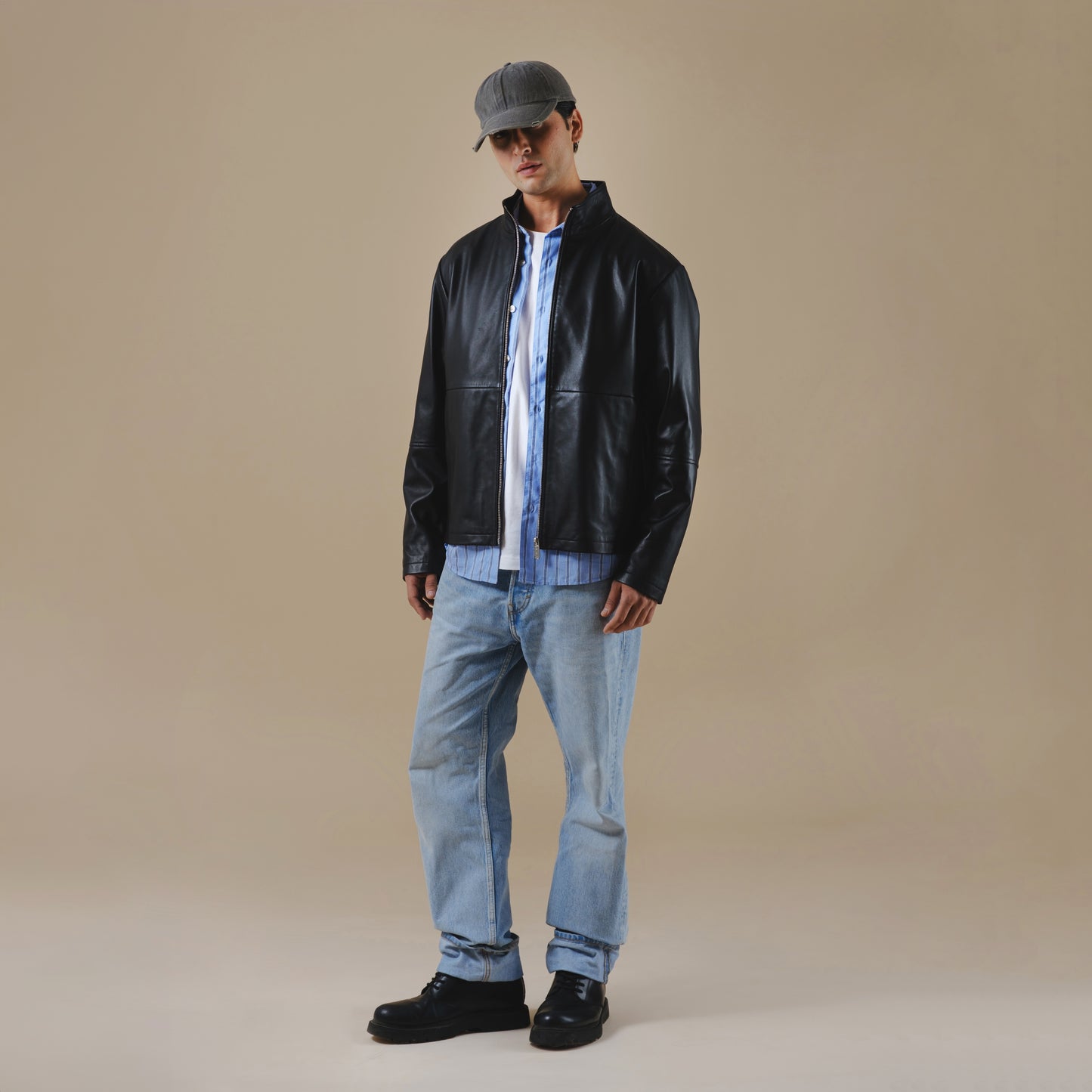 Man wearing a black leather jacket, light blue jeans, and a cap on a beige background